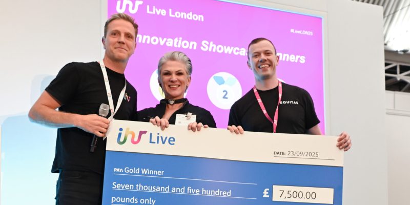 "Three people smiling on stage at IHR Live London 2025, holding a large cheque for £7,500 awarded to the Gold Winner of the Innovation Showcase. A bright pink screen behind them displays the event logo and award title."