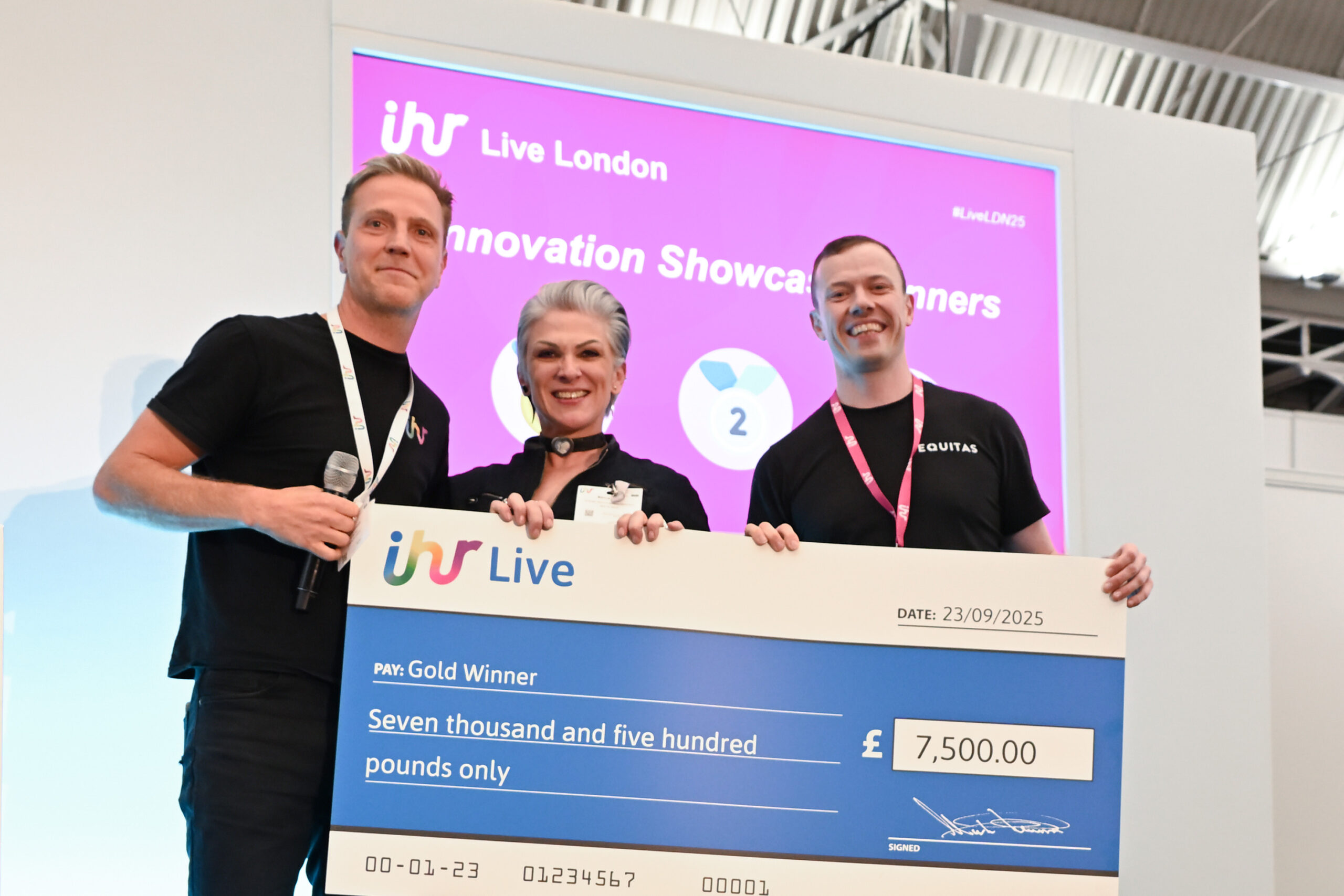 "Three people smiling on stage at IHR Live London 2025, holding a large cheque for £7,500 awarded to the Gold Winner of the Innovation Showcase. A bright pink screen behind them displays the event logo and award title."